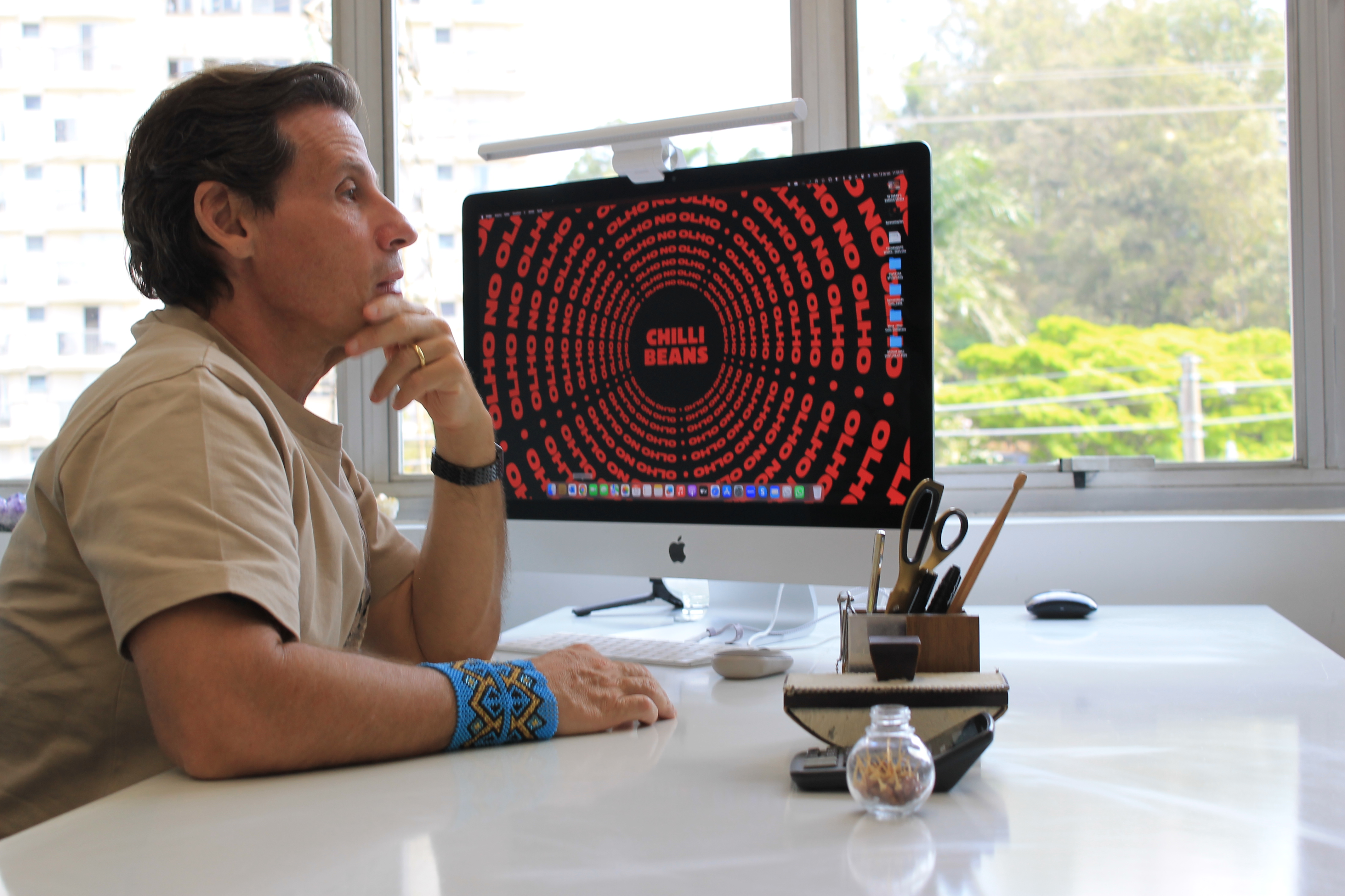 O empresário, de pele branca e cabelos castanhos puxados para trás, descansa o queixo sobre a mão esquerda, com um olhar pensativo. Ao fundo, há uma janela grande e, sobre a mesa, dentre itens de escritório, um monitor da Apple com um protetor de tela da Chilli Beans.