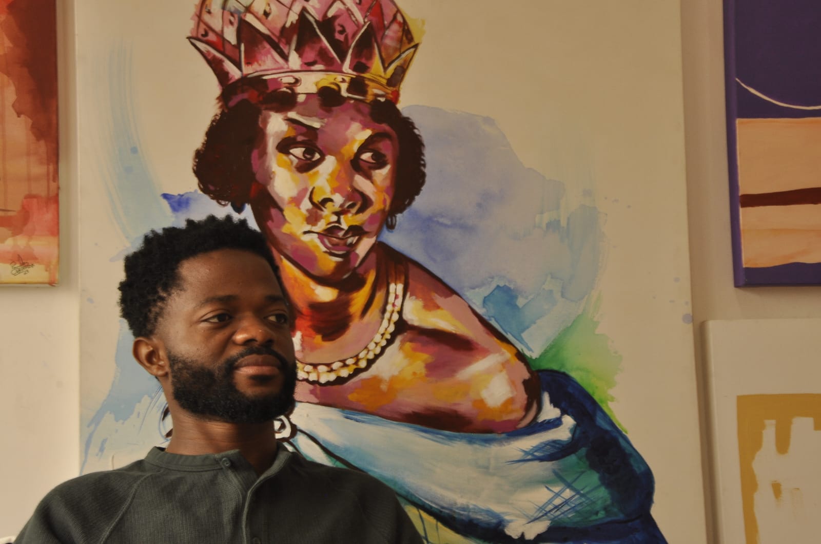 Paulo Chavonga, um homem negro está sentado à frente de uma pintura de uma mulher negra, a rainha Nzinga Mbandi. O homem veste uma camisa cinza de botão. Ele tem barba e cabelos pretos e crespos. Está com uma expressão serena no rosto, com a cabeça levemente virada para a direita. A mulher pintada atrás têm a cabeça pintada na mesma direção. É uma figura da realeza: uma coroa pousa sobre seu cabelo crepso, e um colar dourado envolve seu pescoço. Veste uma roupa de gala azul. O jogo de iluminação da pintura faz parecer que a pele da rainha reluz dourado.