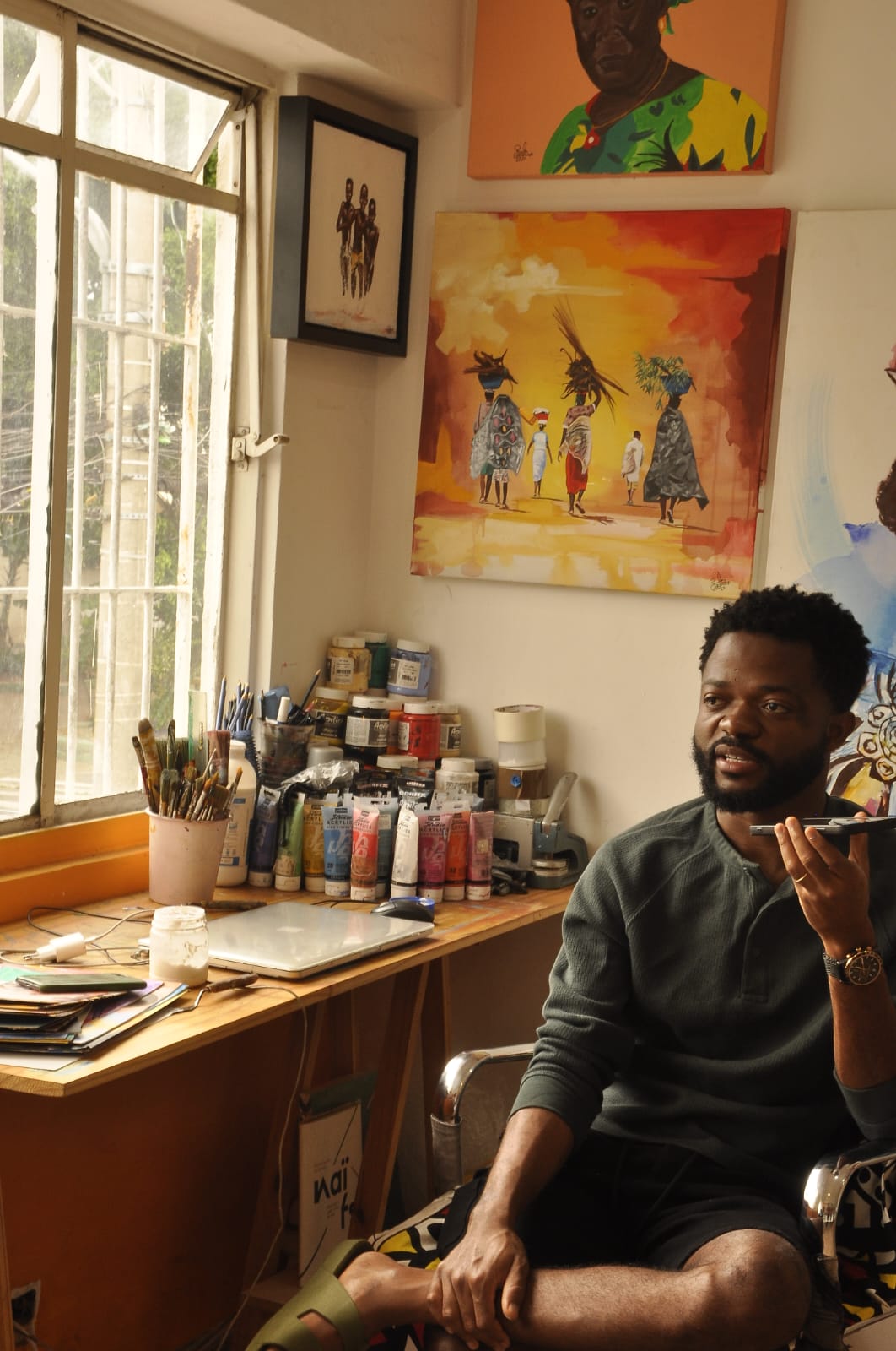 Paulo Chavonga sentado em seu ateliê, com o cotovelo apoiado na cadeira enquanto usa o celular. O ambiente é iluminado pela luz da janela, uma mesa de madeira ocupa o lugar, em cima dela estão alguns utensílios artísticos como tintas, pincéis e um notebook. Dois quadros em tons de amarelo e azul estão pendurados na parede atrás de Paulo.