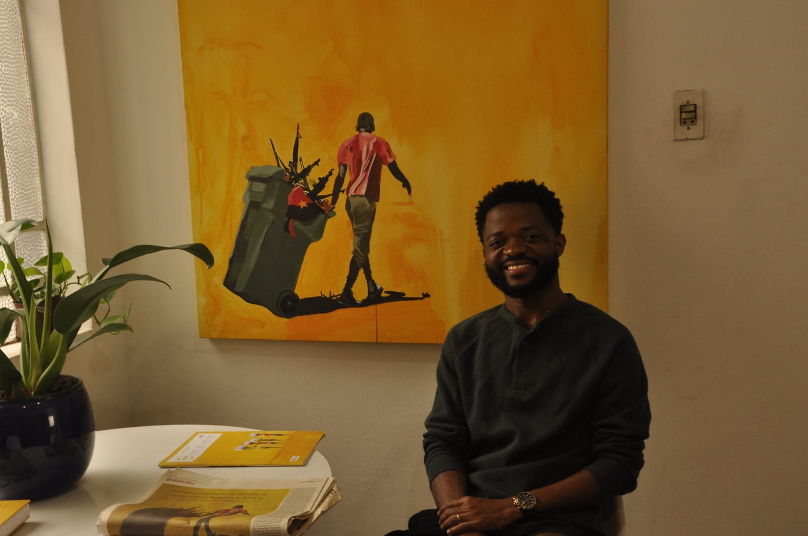 O pintor Paulo Chavonga, um homem negro, está posando sentado. Ao lado dele, uma mesa branca. Sobre ela, um vaso azul escuro com uma planta pequena e verde, um jornal com a foto da sua obra "Não Recicláveis" e um livro amarelo. Atrá de Paulo, a tela da obra "Não Recicláveis", que tem um fundo amarelo, com um homem negro carregando uma ata de lixo. O homem veste uma blusa vermelha, uma calça verde, e um tênis branco. Dentro do lixo verde que carrega, armas de guerra, e uma bandeira prata e vermelha, com uma estrela amarela no centro.  A bandeira lembra a de Angola.