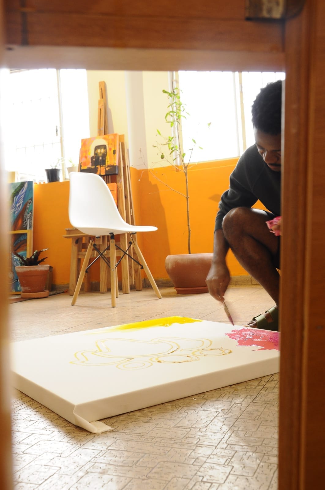 O cenário é um recorte do ateliê de Paulo. O chão tem a cor creme, e sobre ele, uma tela de pintura. A tela tem três o corpo do que parecem ser pessoas desenhadas, contornadas de amarelo. No canto direito inferior da tela, uma mancha amarela. No canto superior direito, uma mancha rosa que está sendo pintada pelo Paulo. Ele está agachado em cócoras, e apoia o braço esquerdo no joelho, com o pincel na mão. De fundo uma cadeira branca, ao lado de um vaso marrom com uma planta pouco cheia. Um cavalete com duas telas está apoiado sobre a parede laranja