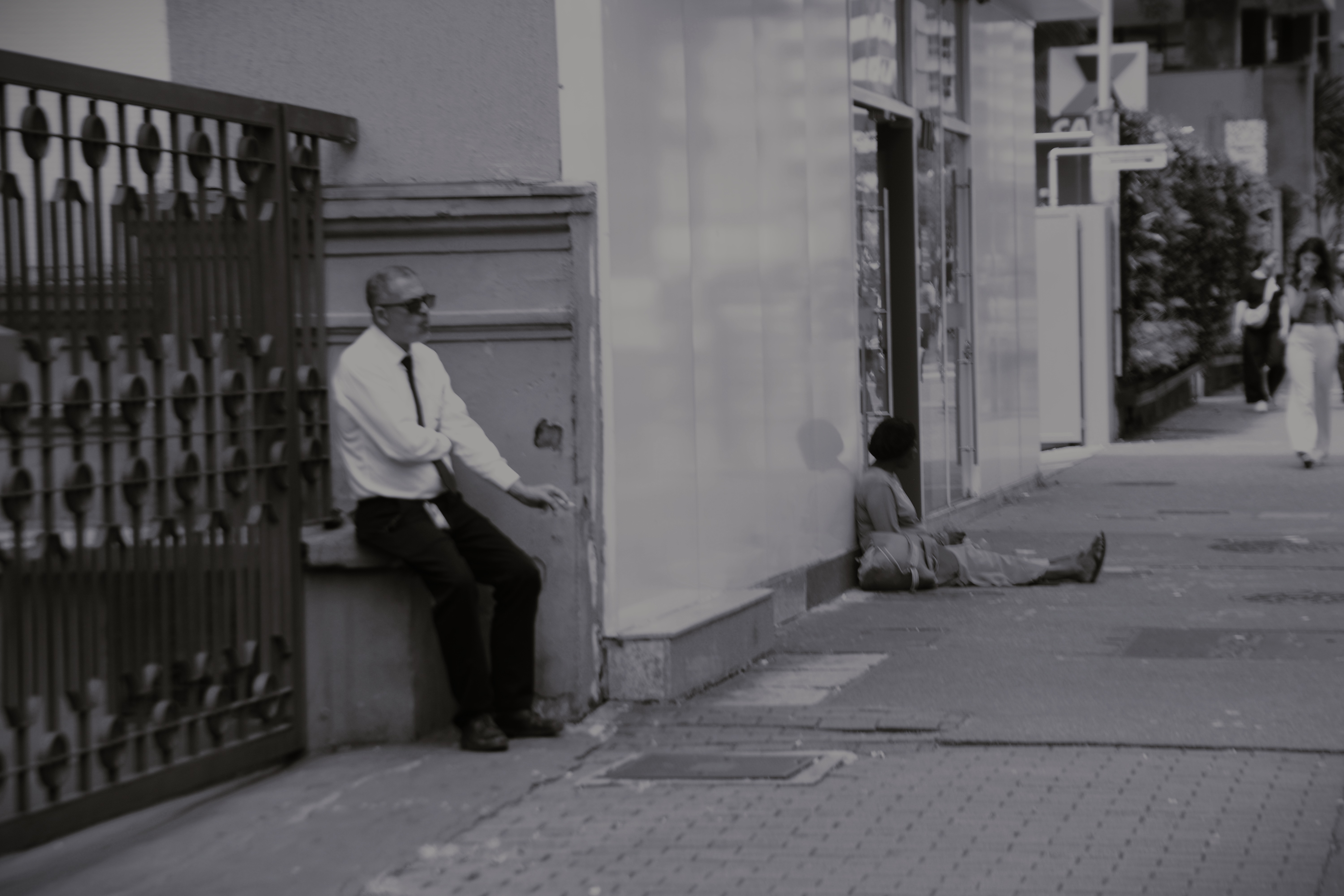 Um trabalhador fuma um cigarro na calçada enquanto uma mulher em situação de rua está sentada ao fundo - foto: Heitor Landim / Agenzia