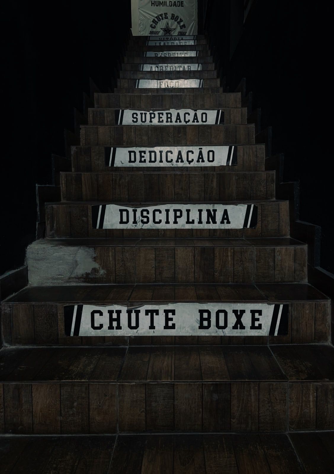 Escadaria de madeira em que cada degrau possui uma faixa branca com palavras em preto, como "Chute Boxe", "Disciplina", "Dedicação" e "Superação", servindo como guia motivacional no ambiente.
