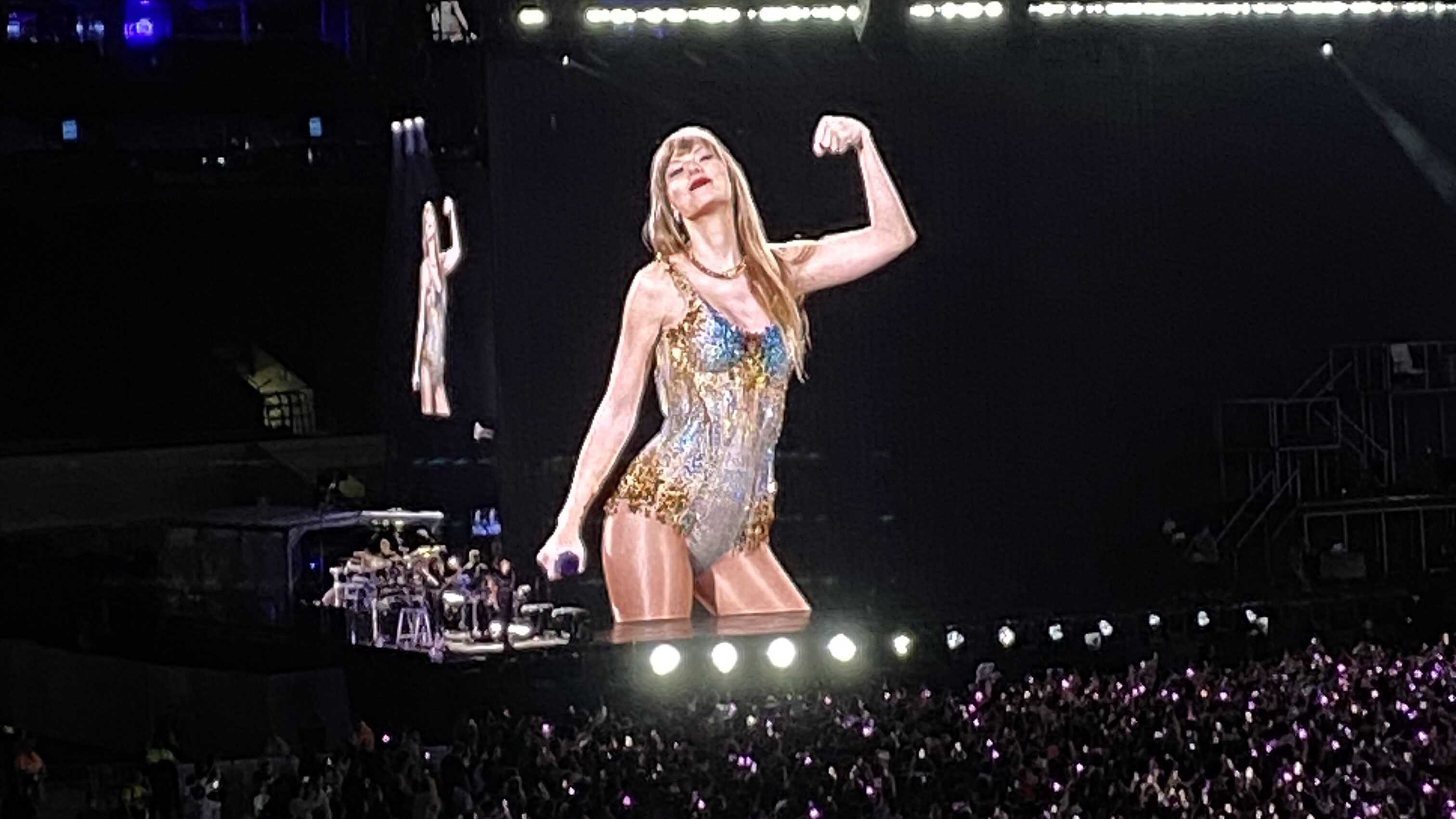 Estádio lotado no show da cantora Taylor Swift em São Paulo, as luzes estão apagadas com apenas um foco de luz sobre a cantora, que é loira, branca e veste uma roupa multicolorida e brilhante. A plateia está lotada com lanternas de celular ligadas irradiando vários pontos de luz no estádio escuro. 
