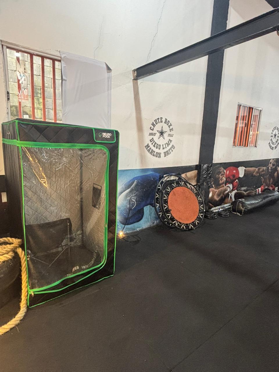 Cabine de sauna portátil preta com detalhes em verde neon. No interior, há uma cadeira dobrável simples posicionada para uso.