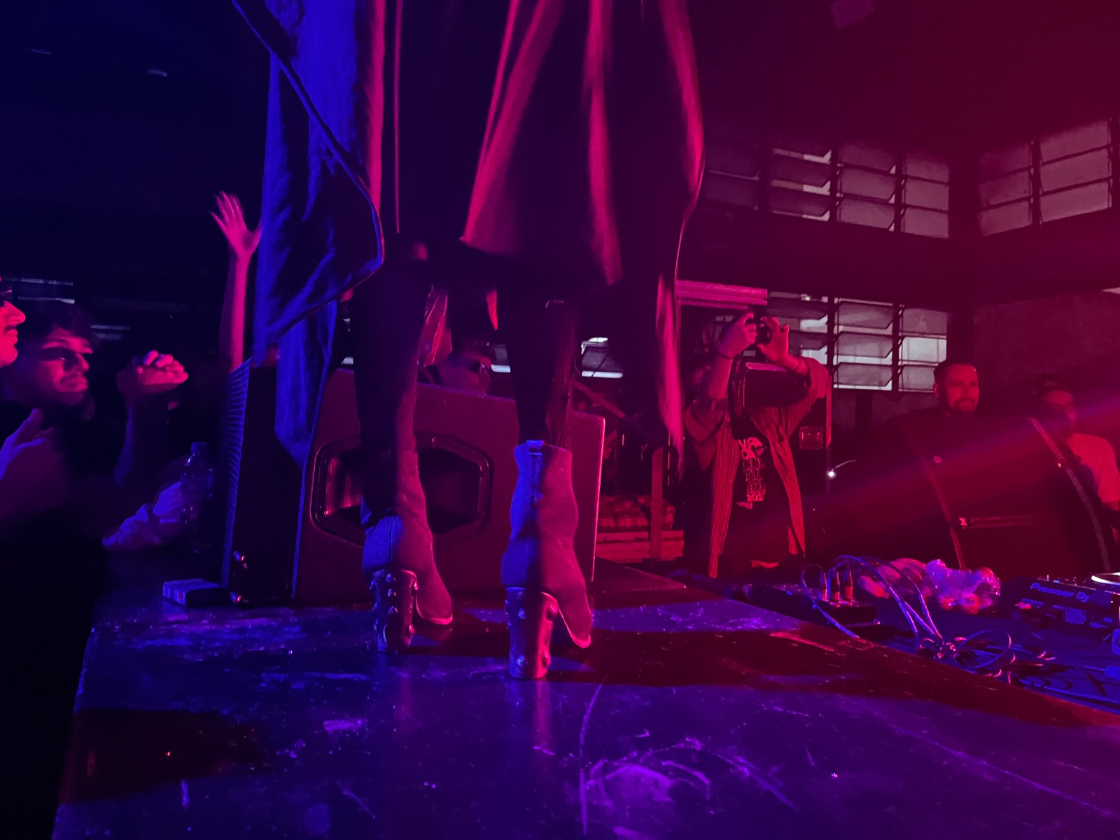 A foto mostra a parte inferior de uma pessoa usando botas de salto alto com detalhes metálicos, em um palco iluminado por luzes roxas e vermelhas. Em volta do palco, várias pessoas observam e algumas levantam as mãos, enquanto uma pessoa segura uma câmera para registrar o momento. O ambiente tem um aspecto industrial, com janelas grandes ao fundo.