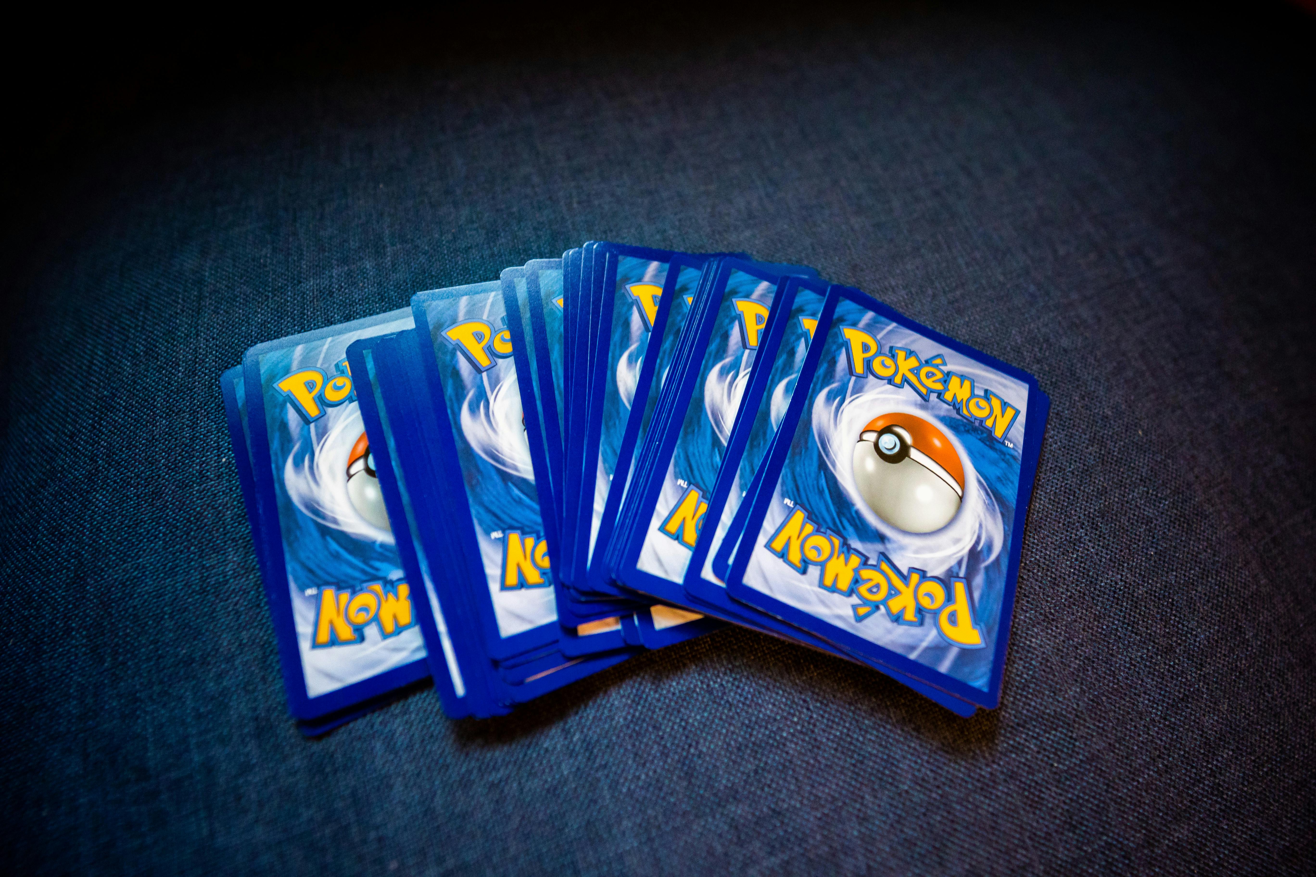 Imagem de várias cartas do Pokémon Trading Card Game, derivado da série de videogames Pokémon, dispostas em leque sobre uma superfície escura de tecido. As cartas estão viradas para baixo, mostrando o verso azul com uma esfera central (Poké Bola) e o nome “Pokémon” em amarelo. A iluminação destaca o centro da imagem, enquanto as bordas ficam mais escuras, criando um efeito de foco nas cartas. - Caleb Oqueno