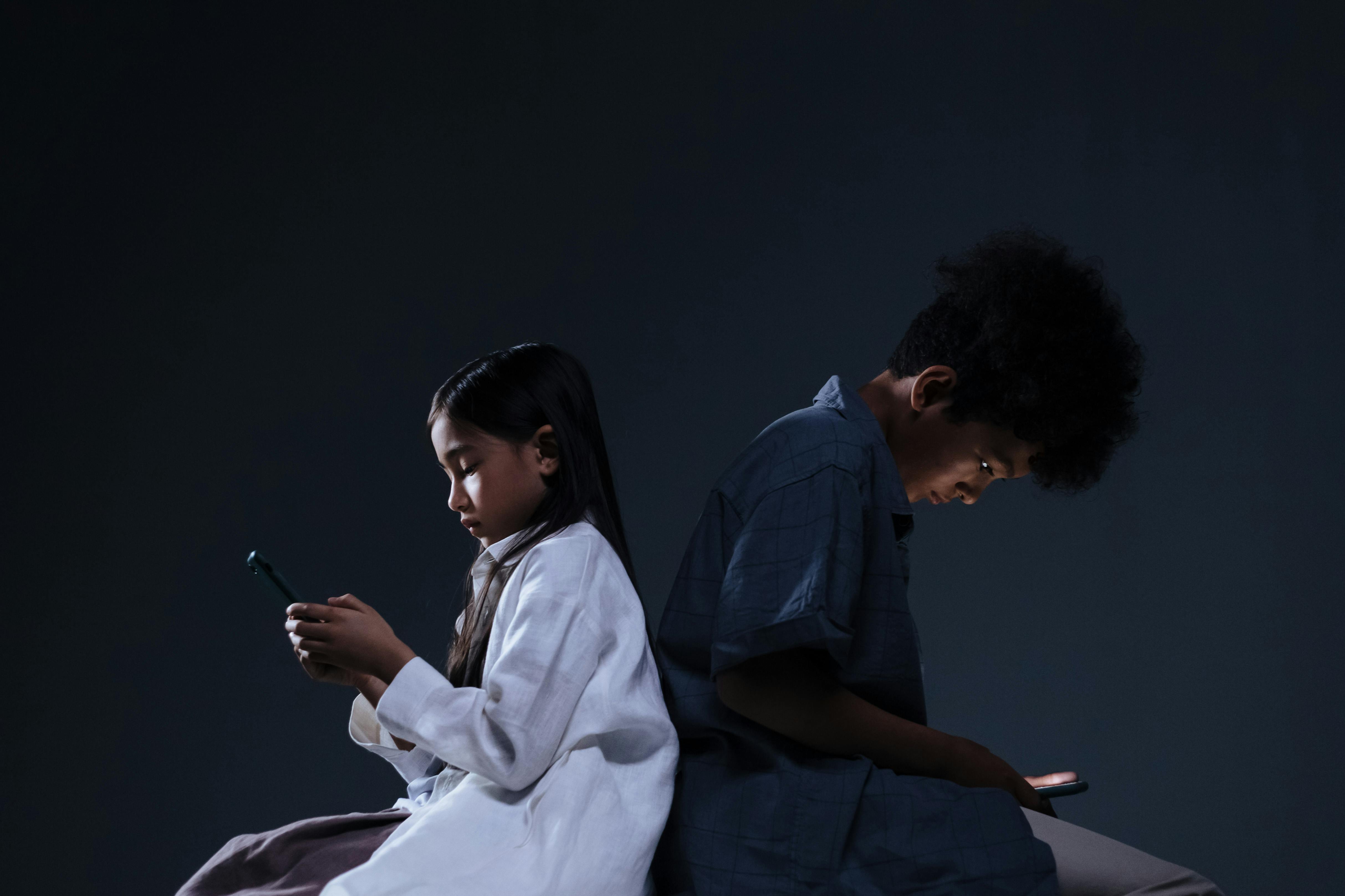 A imagem apresenta duas crianças, um menino e uma menina, de costas um para o outro mexendo em seus respectivos telefones.